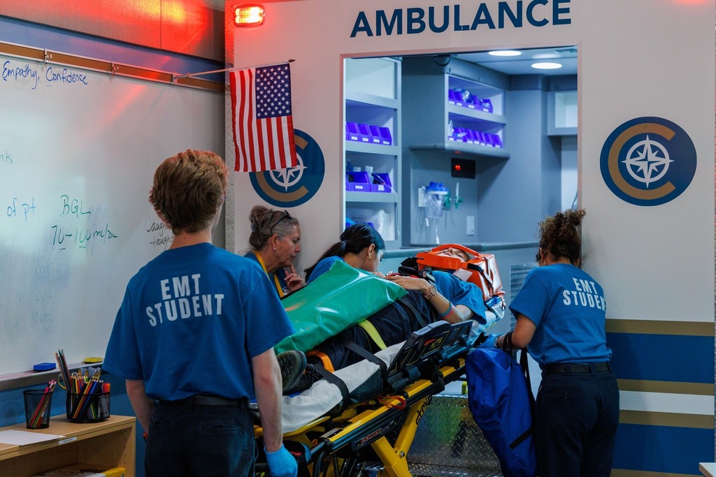 EMT students guide the stretcher into a simulated ambulance bay during training, practicing real-world patient transfer procedures.
