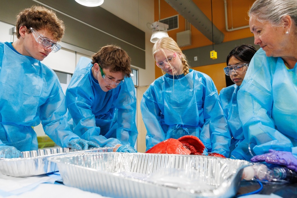 A group of EMT students in blue gowns and goggles gathers around trays containing anatomical training materials, closely examining and learning with their instructor.