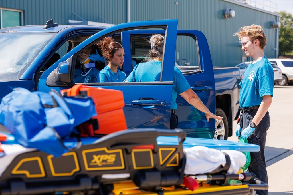 : EMT students in blue uniforms assist with a training scenario involving a patient in a blue truck, preparing equipment and a stretcher for extraction.