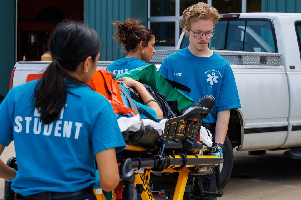 A team of EMT students wheels the secured patient on a stretcher toward the ambulance area, continuing simulated emergency care.