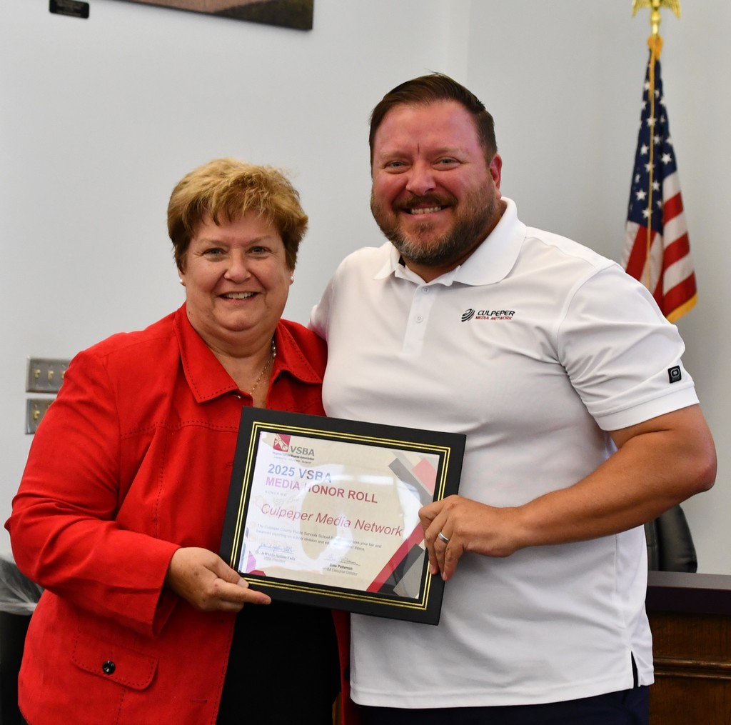 A Culpeper County School Board member presents a framed 2025 VSBA Media Honor Roll certificate to a representative from Culpeper Media Network. Both are smiling and standing together in front of an American flag.