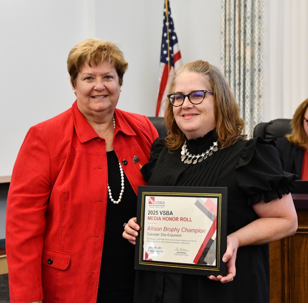 A Culpeper County School Board member presents a framed 2025 VSBA Media Honor Roll certificate to a representative from the Culpeper Star Exponent. Both are smiling and standing together in front of an American flag.