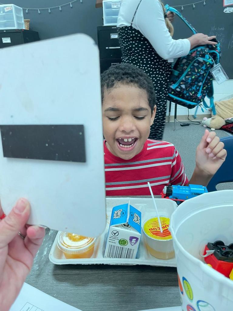 Student in a red striped shirt laughing while a whiteboard is held up in front, with a lunch tray and milk carton on the desk.