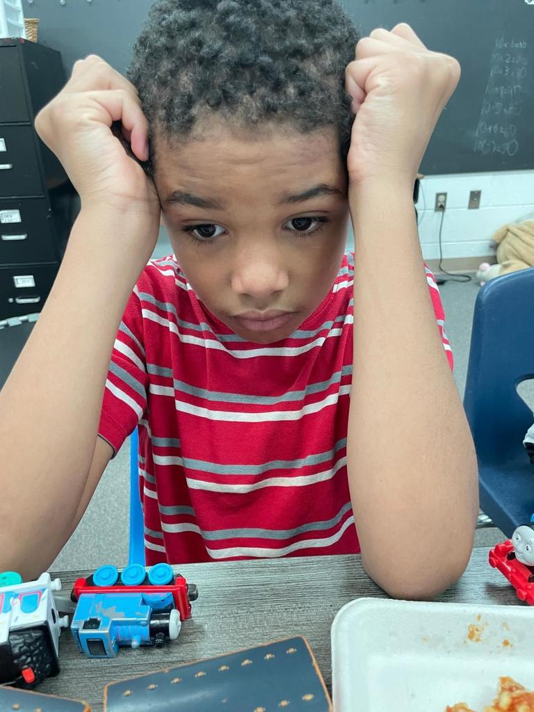Student in a red striped shirt resting head in hands at a table with toy trains and a lunch tray.