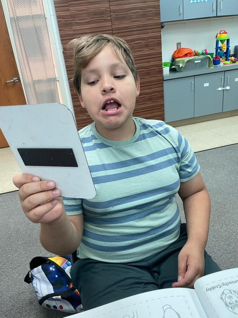 Student in a striped shirt holding up a whiteboard while practicing sounds, mouth open mid-speech.