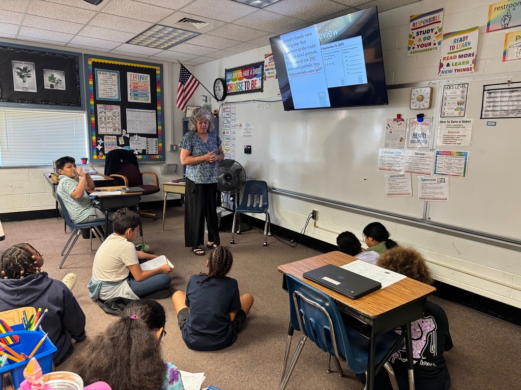 A classroom teacher, Mrs. Dodson, stands at the front of the room leading a math lesson displayed on a large screen while students sit on the carpet and at desks, listening and writing in notebooks.