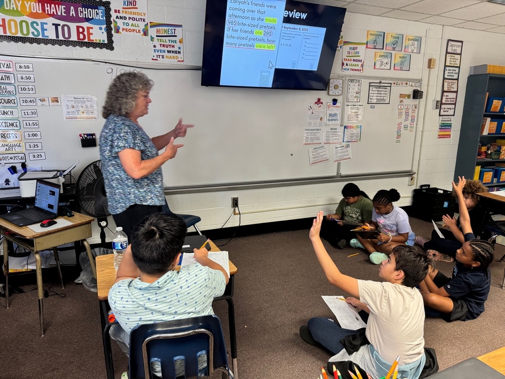 Mrs. Dodson leads a math review in front of the class. Several students sitting on the carpet have their hands raised to answer a question displayed on the screen.