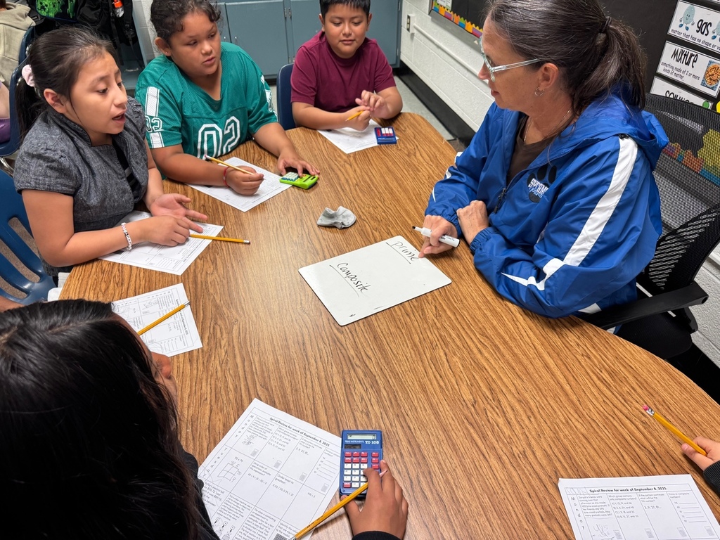 Mrs. Carter sits at a round table with a small group of students. They are working on math problems using worksheets and calculators, with Mrs. Carter writing on a small whiteboard.