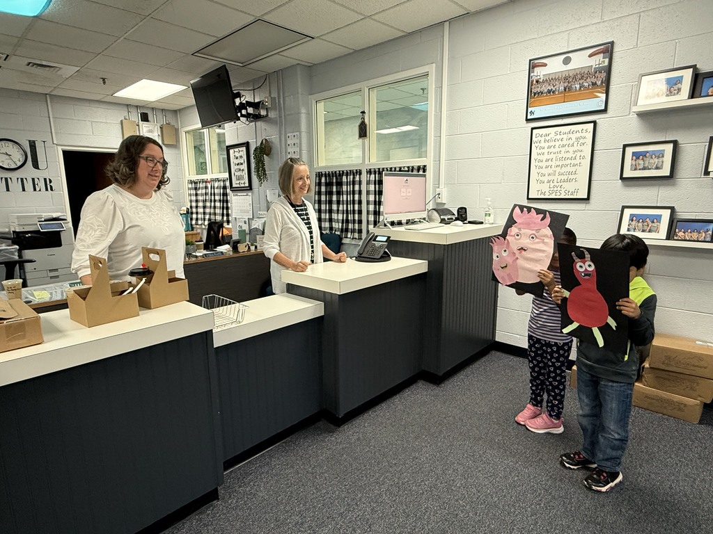 The two children display their monster artwork inside a school office, standing in front of a counter where two smiling staff members watch. The staff appear amused and encouraging as the children hold up their large pink and red monster creations.