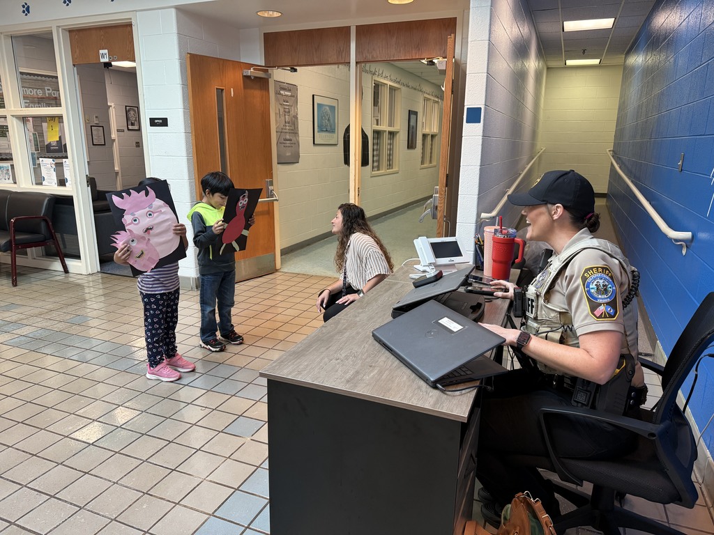 The same two children stand in a school lobby showing their monster artwork to a sheriff’s deputy seated at a desk. The deputy smiles and talks with them while another adult kneels nearby, engaging with the students.