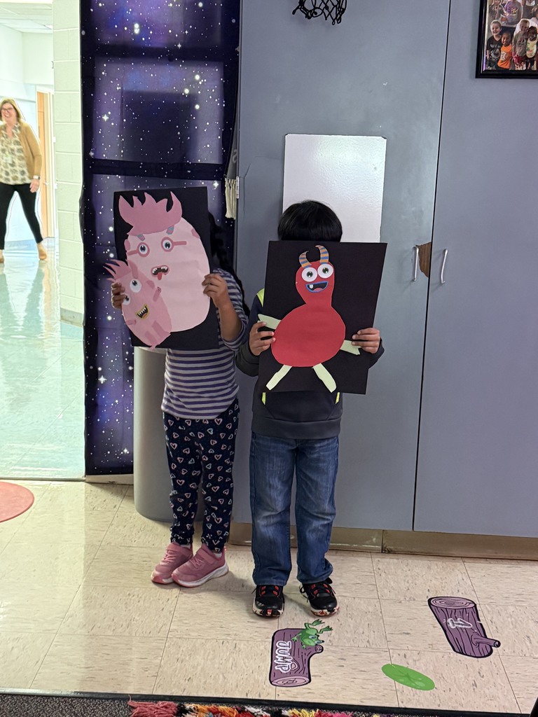 Two children stand side by side in a hallway, each holding up large black construction paper with colorful monster artwork covering their faces. The child on the left holds a pink monster with big eyes, teeth, and spiky hair, while the child on the right holds a red monster with striped horns, wide eyes, and green legs.