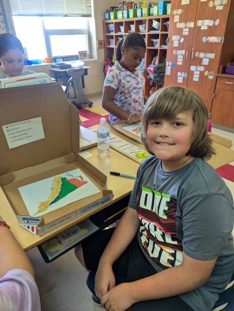 A boy smiles at the camera while sitting at a desk with a clay Virginia map in front of him. Two classmates behind him work on their own projects.