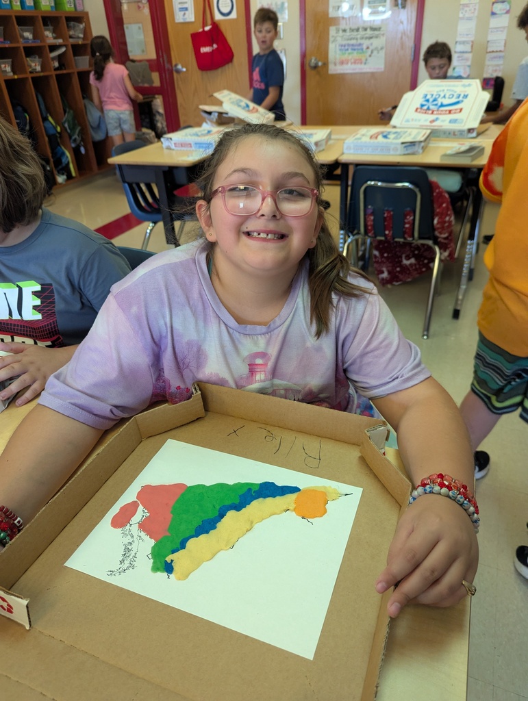 A student with glasses smiles at the camera, leaning on her desk with a finished clay Virginia map in front of her. The map shows distinct colored regions made with clay inside a pizza box tray.