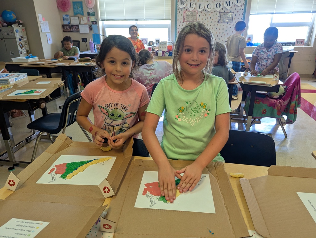 Two smiling students sit at desks working on clay models of Virginia’s regions. Their maps are placed inside recycled pizza boxes to keep the projects contained. Other classmates work in the background.