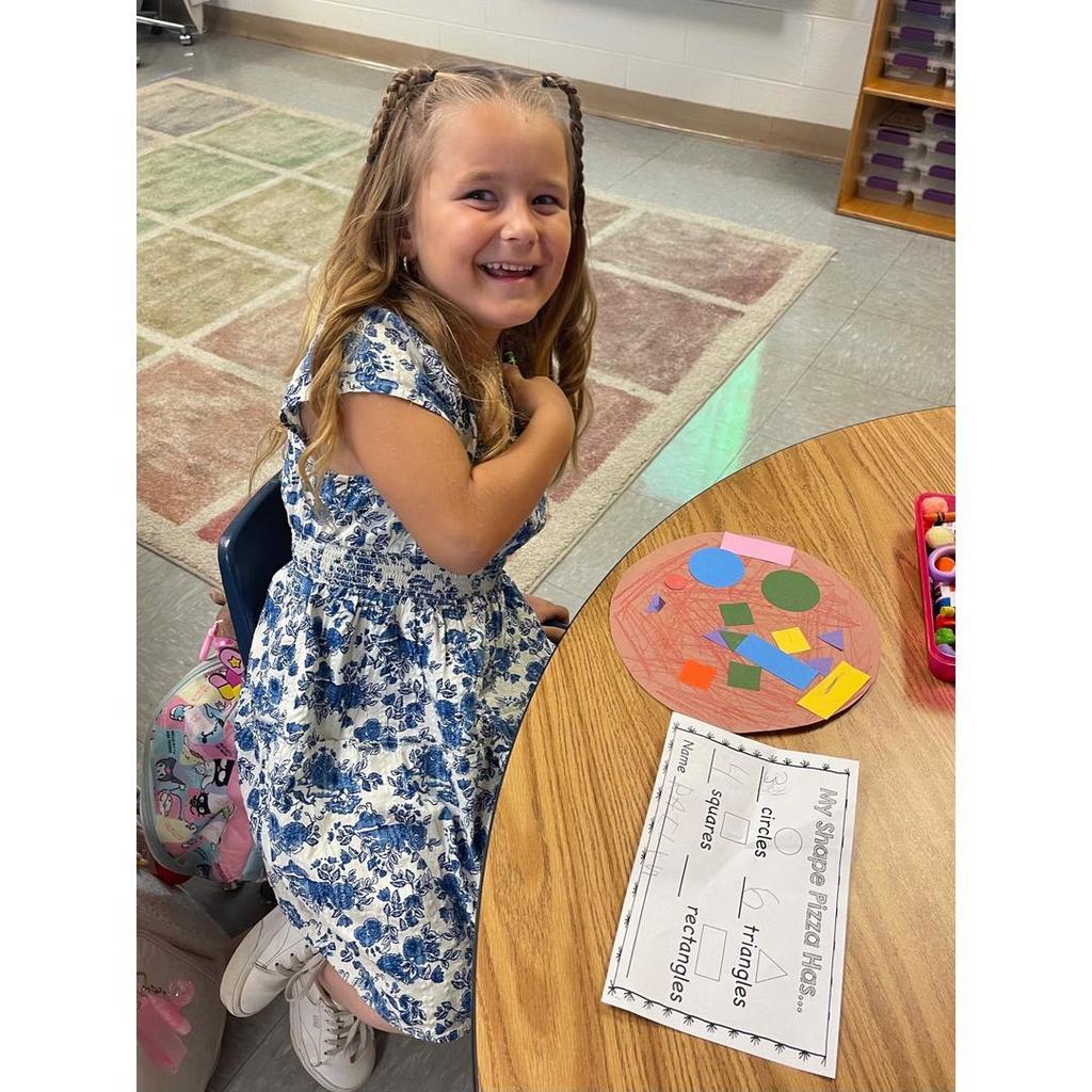 A smiling student in a blue-and-white dress sits at her desk with a shape pizza and a worksheet titled “My Shape Pizza Has.”