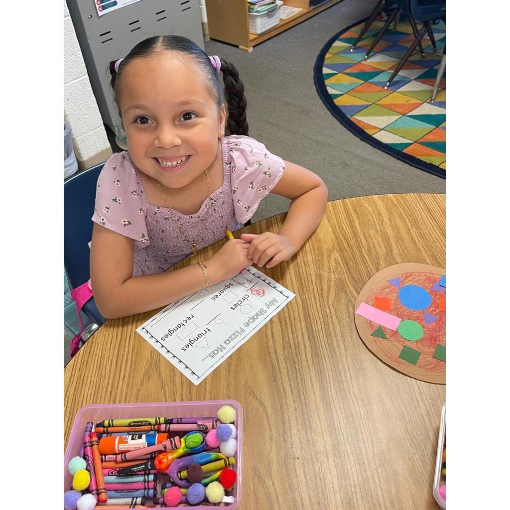 A student in a light purple top smiles with her shape pizza and worksheet on the table.