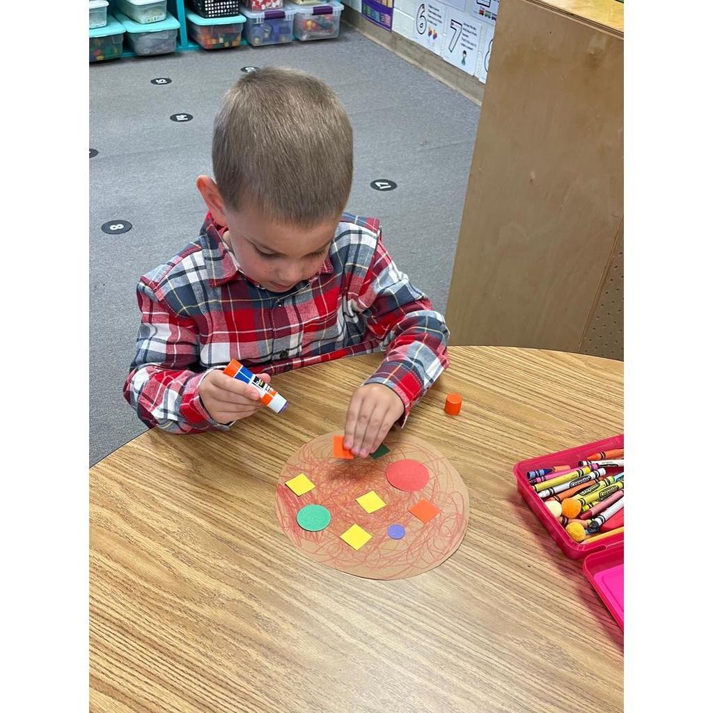 A student in a red plaid shirt concentrates on gluing shapes onto his paper pizza