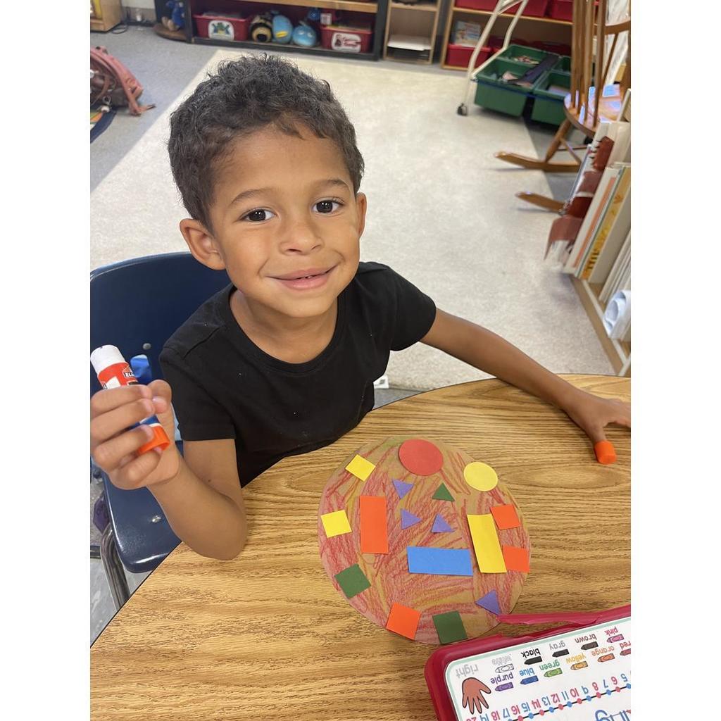 A student in a black shirt proudly shows his glue stick and shape pizza covered in colorful shapes