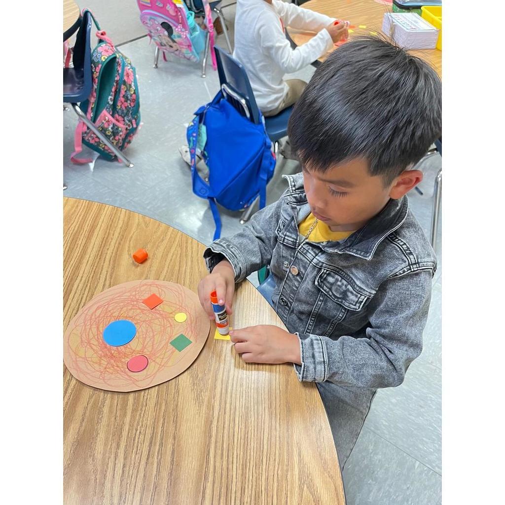 A student glues a yellow square onto a paper pizza decorated with circles, squares, and other shapes.