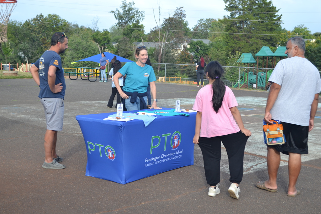 Image contains a table on the playground "PTO Farmington Elementary School" with a volunteer talking to another student and parent.