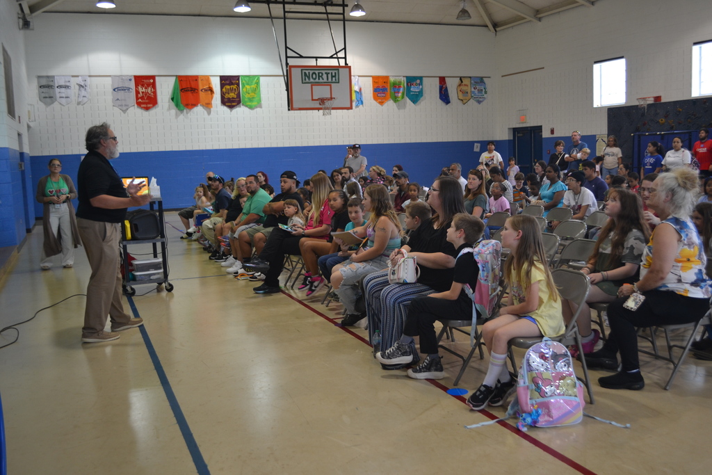 Image contains students and parents sitting in seats in the school gymnasium while the principal presents at Back to School night.