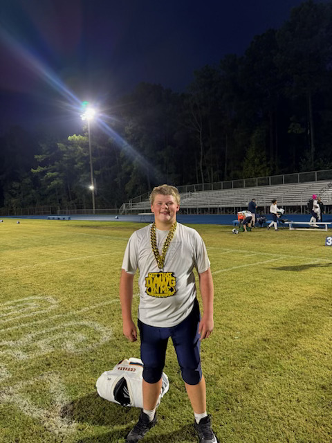 A smiling football player stands on a lit football field at night, wearing practice gear and a large gold “Savage Dawgs” chain. His helmet rests on the grass in front of him, with empty bleachers and trees visible in the background.