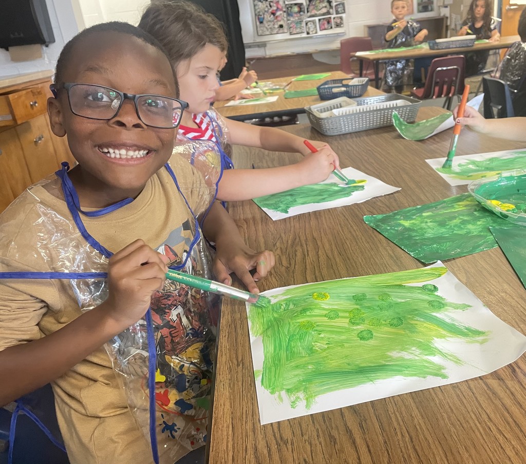 Image contains a  smiling student while painting green artwork.  Other students are in the background working too.