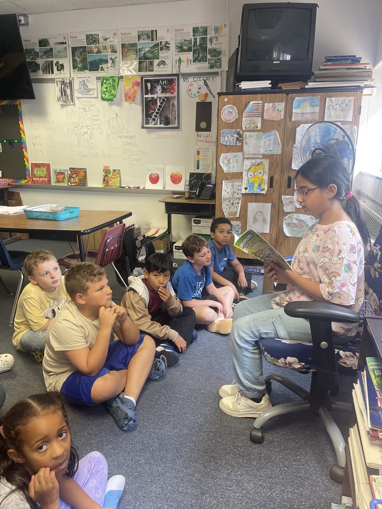 Image contains a student reading a book to her classmates.