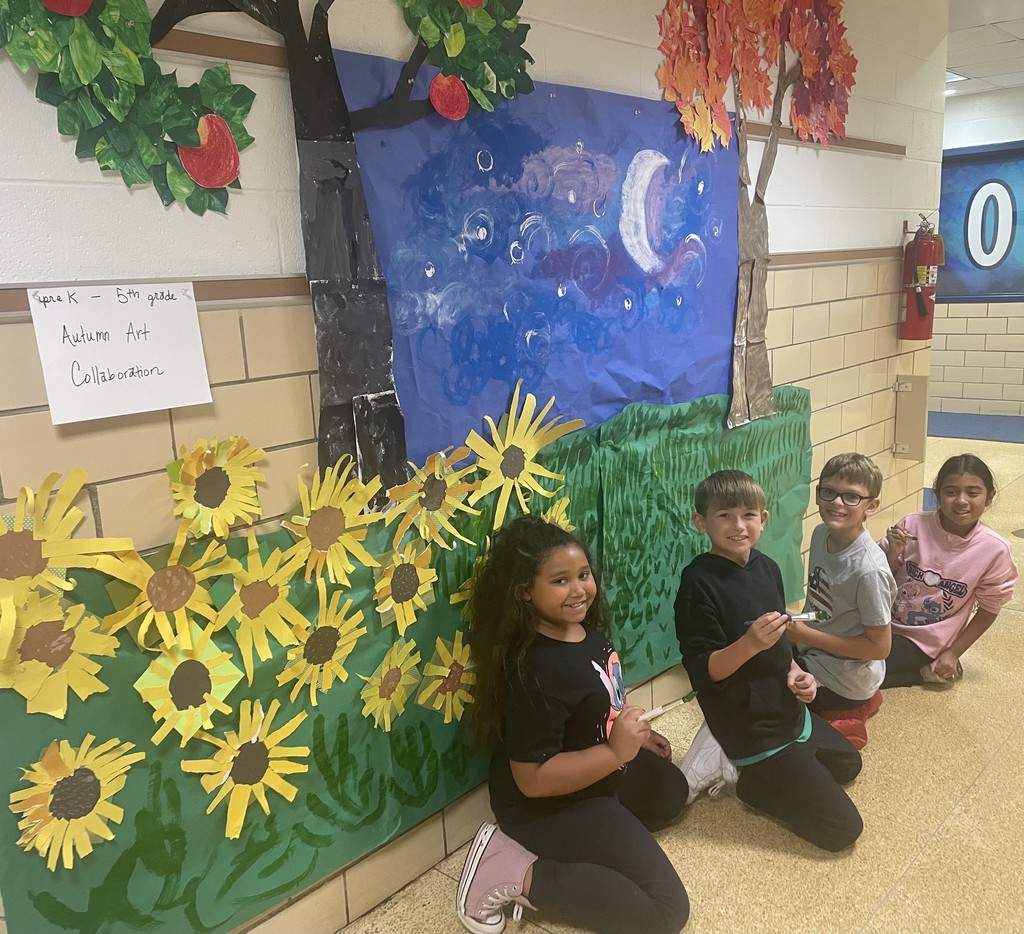 Image contains 4 students bent down adding their artwork to a collage in the school hallway.