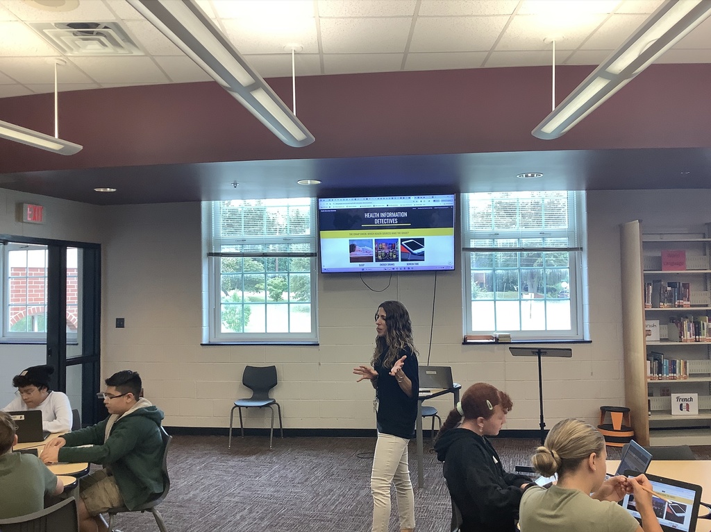 A classroom or library setting with students seated at round tables working on laptops. A teacher stands at the front near a screen displaying a presentation. The wall has large word art featuring terms like “library,” “create,” “inquire,” and “research.