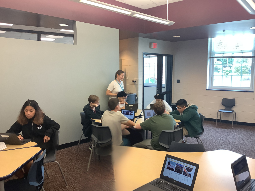 A group of students working together at tables in a library or classroom. Some students are on laptops while a teacher stands nearby assisting. A girl sits separately, working independently on her laptop.