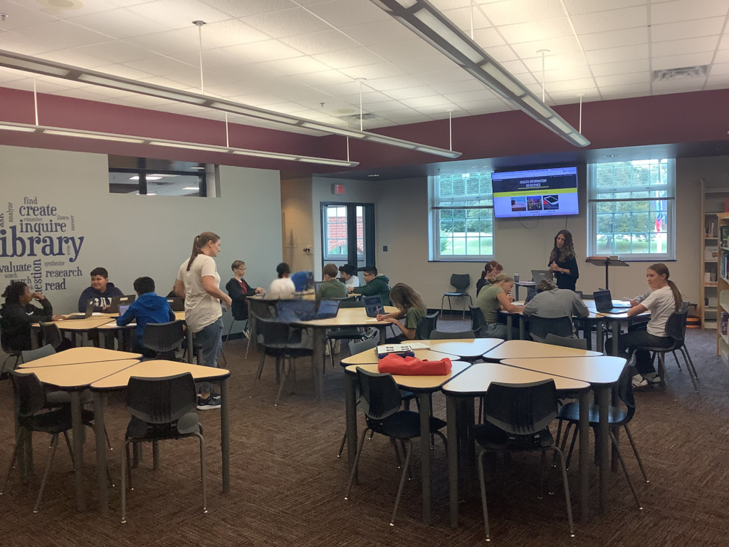 Students are seated at several tables in a library, working on laptops while a teacher gives instructions from the front. A large screen displays a presentation with images and text, and natural light comes through tall windows.