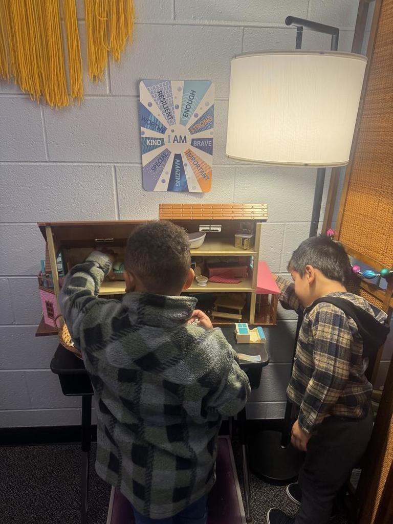 Two boys in plaid and fleece jackets play with a wooden dollhouse under a classroom poster with affirmations like “I am strong” and “I am kind.”