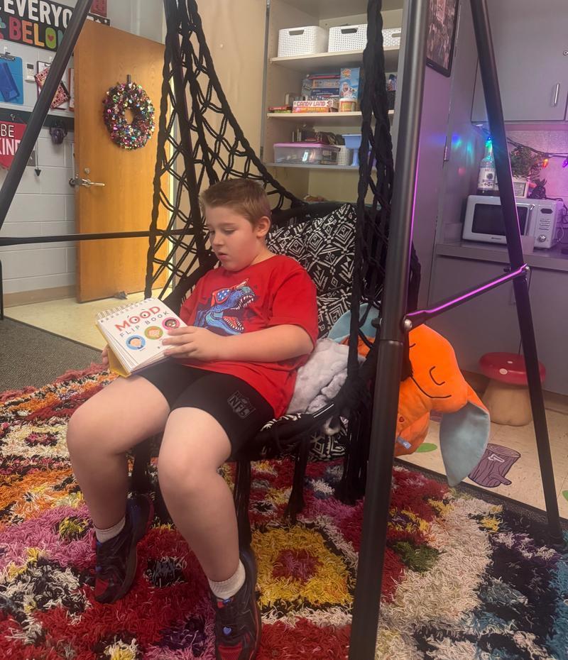A boy in a red shirt sits in a black hanging chair reading a “Mood Flip Book” in a cozy, colorful classroom corner.