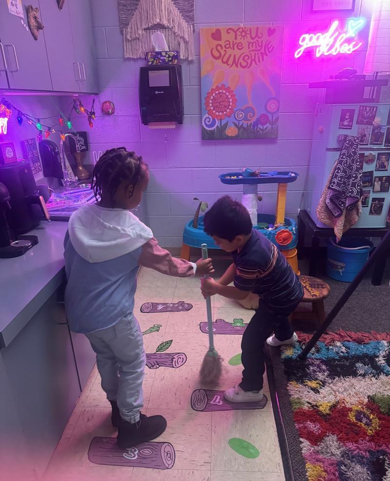 Two children work together with a broom in a classroom play area, surrounded by colorful lights and decorations, under a poster that says “You are my Sunshine.”