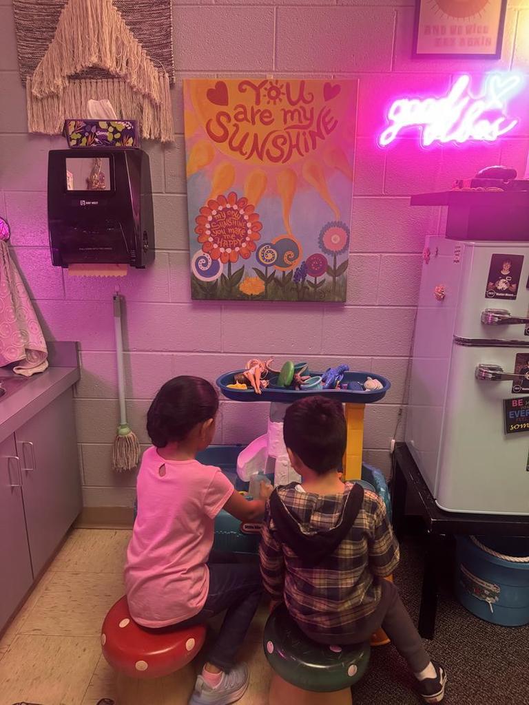 A girl in a pink shirt and a boy in a plaid hoodie sit on mushroom-shaped stools playing together at a toy kitchen set, near a wall decorated with a “You are my Sunshine” poster and a neon “good vibes” sign.