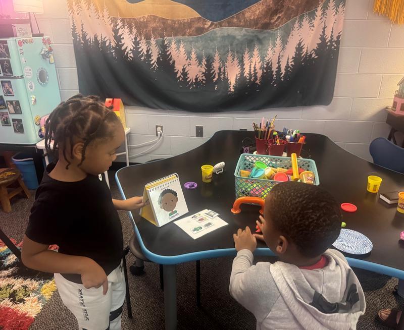 Two children stand at a table with Play-Doh and a mood flip chart, exploring emotions in a creative learning space.