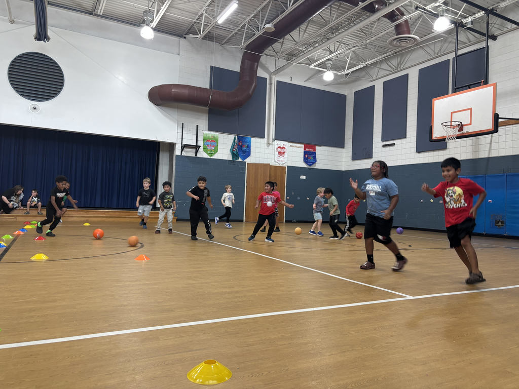 Elementary school students are playing a game in a gymnasium. Several children are running toward balls placed on the floor between colorful cones, while others watch or wait for their turn. The gym has a basketball hoop, stage with a dark blue curtain, banners on the walls, and bright overhead lighting. The activity looks energetic and engaging, with students smiling and moving quickly.