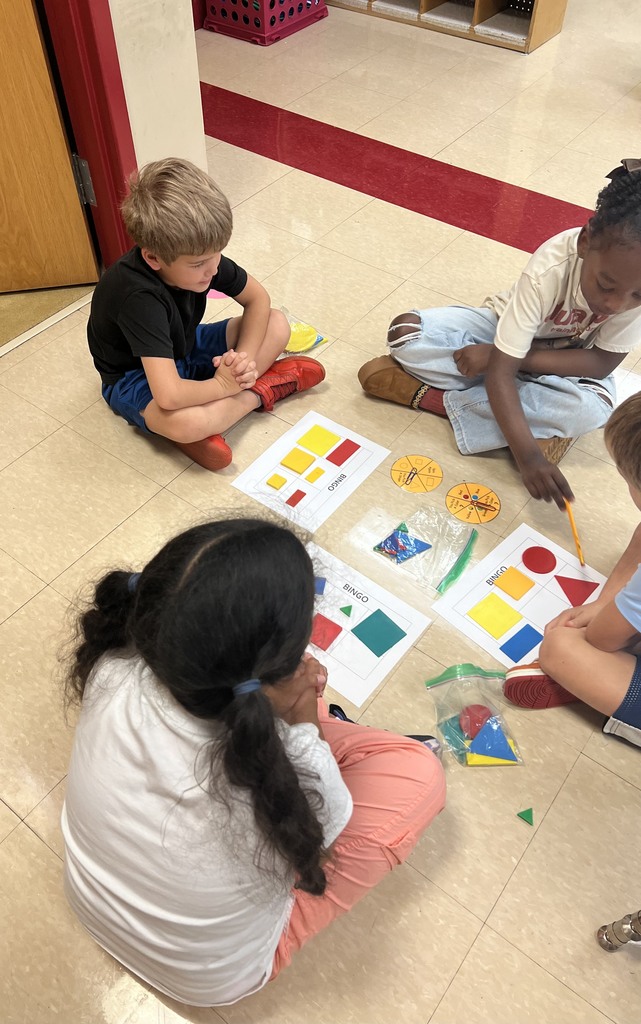 A small group of children sit in a circle on the classroom floor, focusing on bingo cards with bright geometric shapes and a yellow spinner.