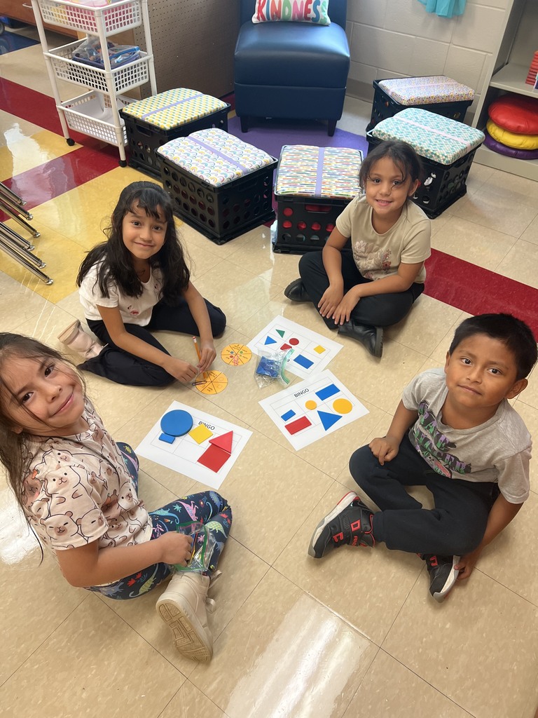 Four young students sit on the floor smiling around bingo boards covered with colorful shapes. A “Kindness” pillow rests on a chair in the background.