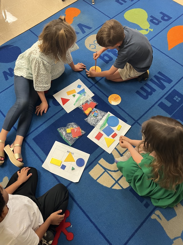 A group of children sit on a blue rug, leaning in toward bingo boards with colorful shapes while one student spins a pencil on a yellow spinner.
