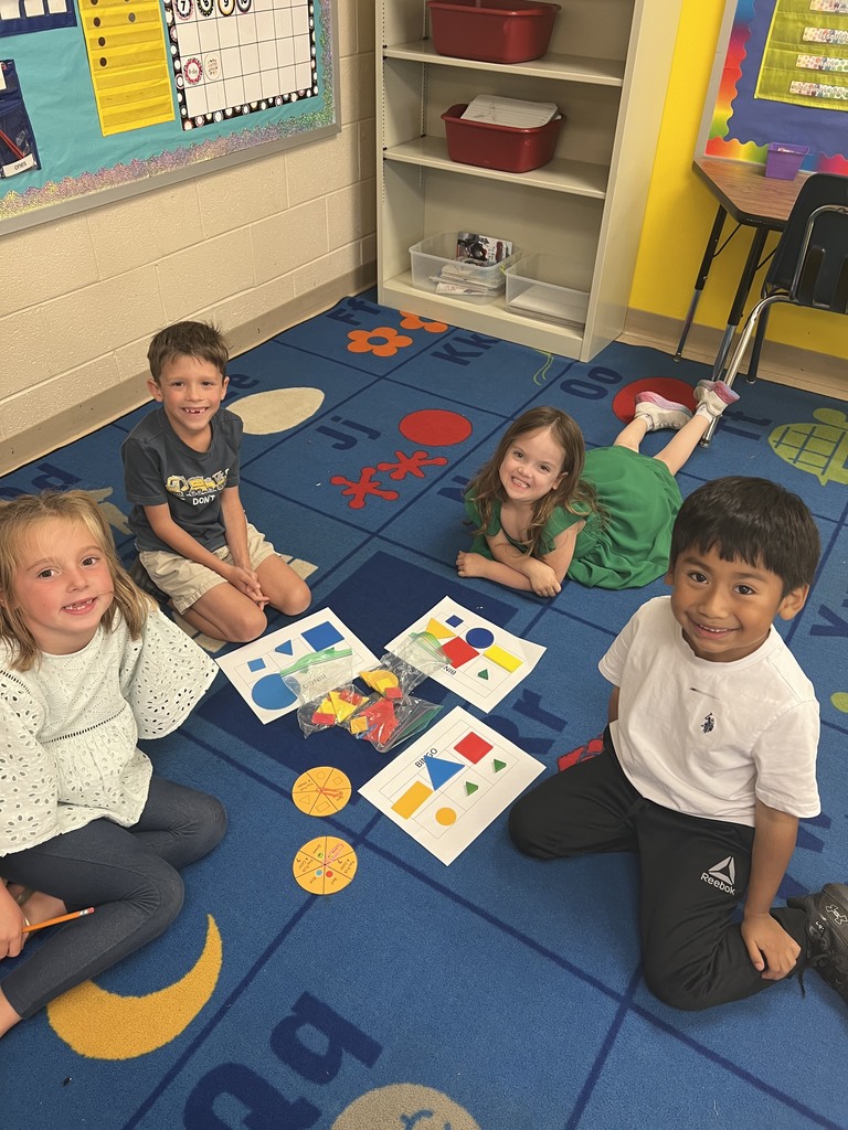 Four children sit together on a blue classroom carpet with bingo boards, shape pieces, and spinners spread out between them.