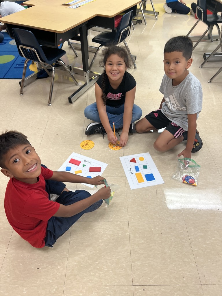 Three students play shape bingo on the floor, grinning as they hold game pieces and pencils.