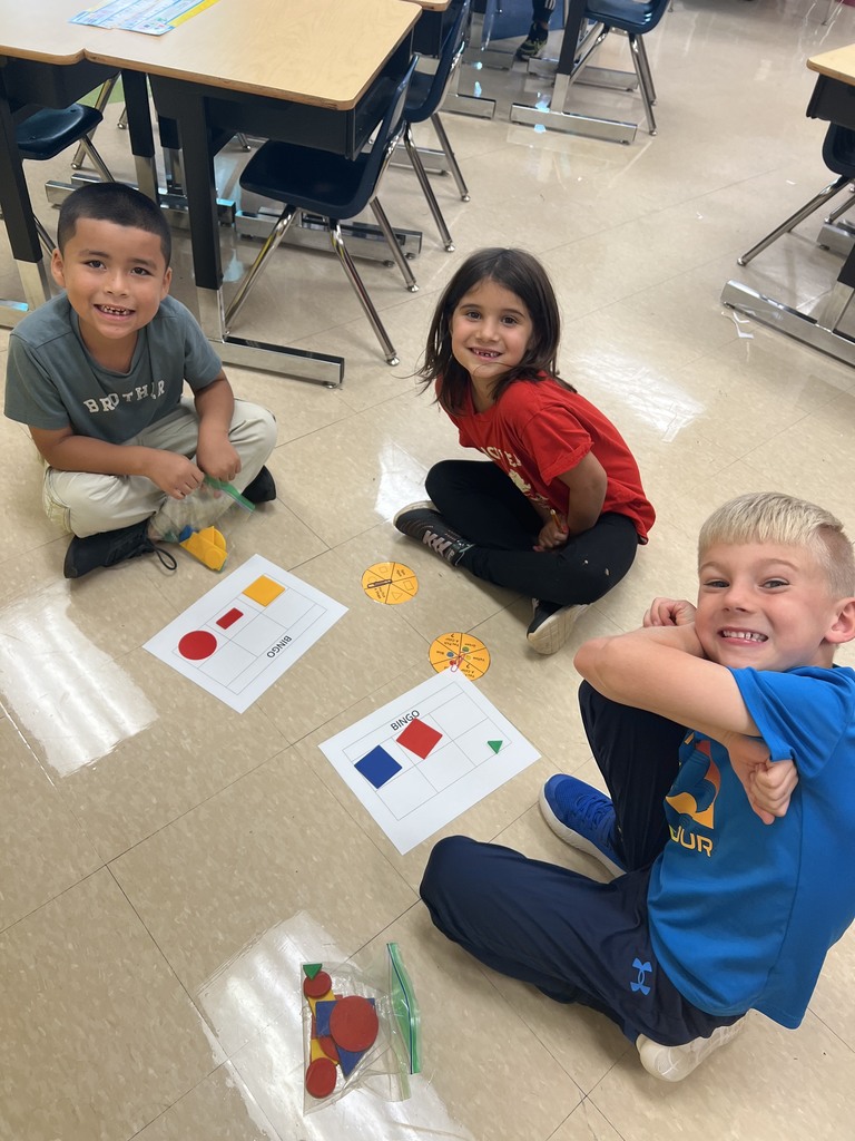Three students smile at the camera while playing a shape bingo game on the floor with laminated boards and plastic pieces.