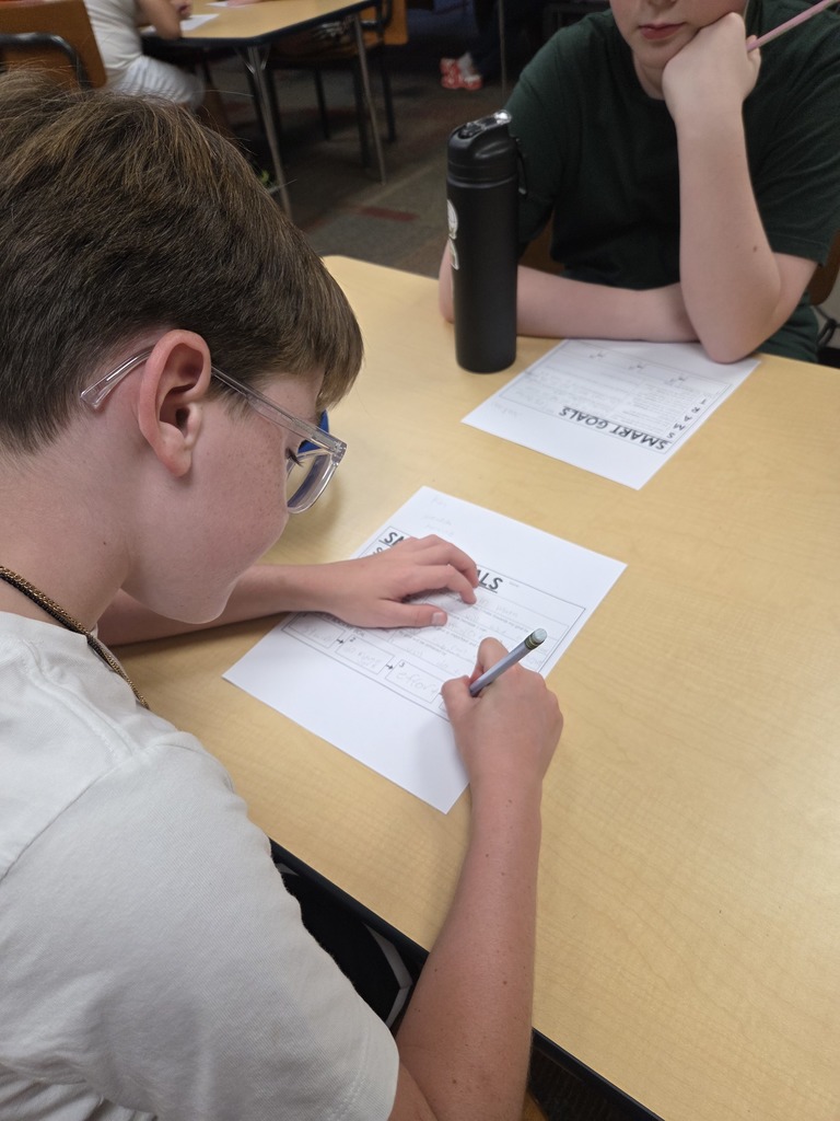 A student wearing glasses leans over their SMART Goals worksheet, writing with a pencil, while a classmate sits beside them at the table.