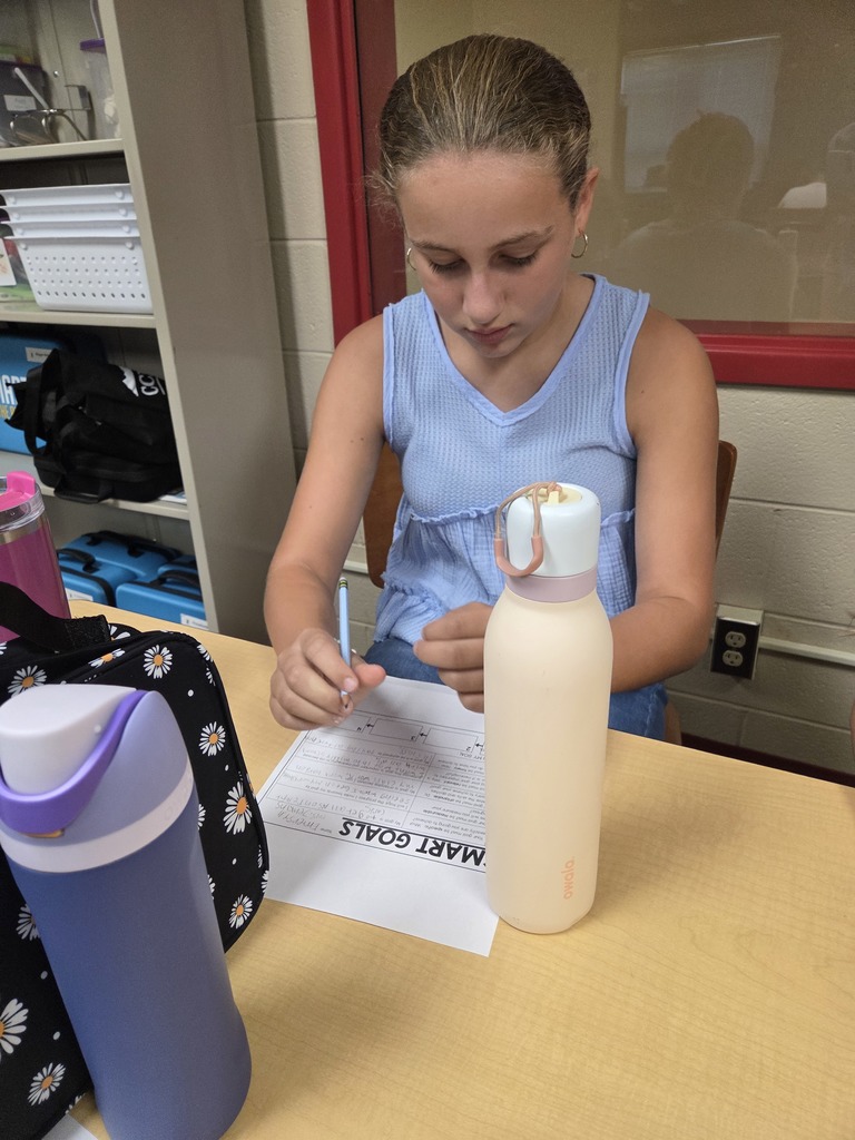 A student in a blue sleeveless shirt sits at a desk, focused on writing their SMART Goals worksheet with water bottles and supplies nearby.