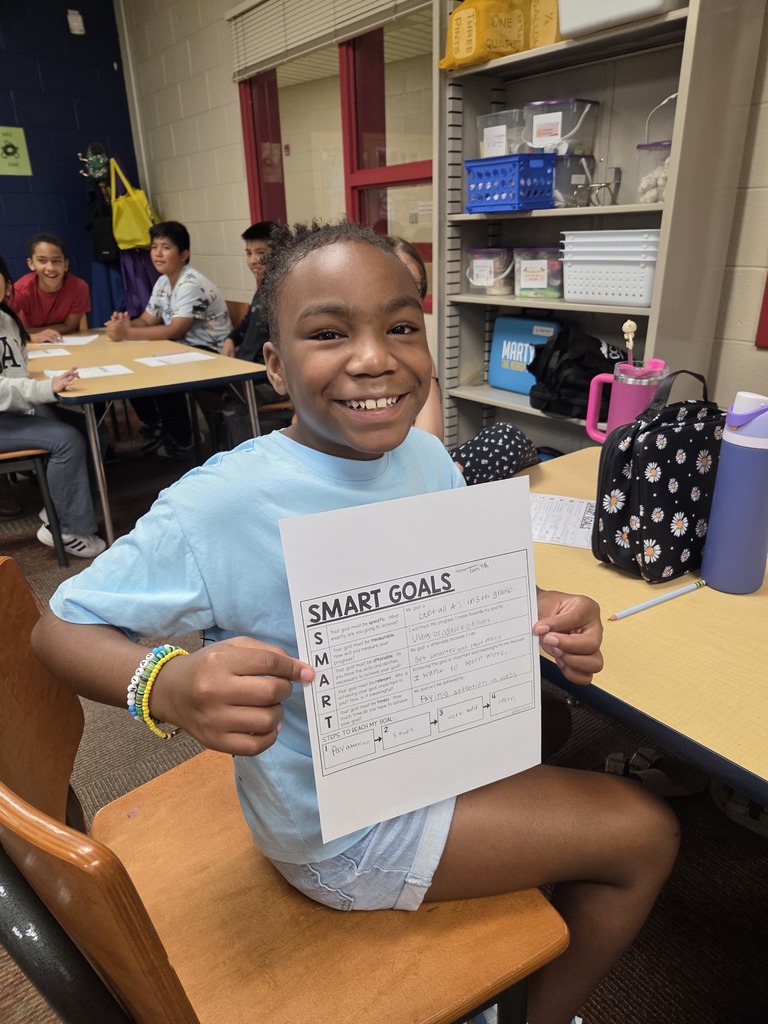 A smiling student holds up their SMART Goals worksheet while sitting at a classroom table. Other students are working in the background.