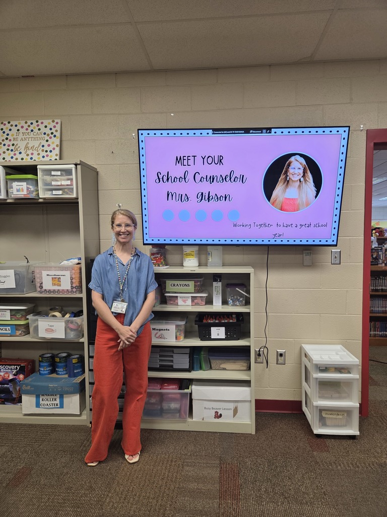 A school counselor stands smiling in front of a large display screen that reads “Meet Your School Counselor Mrs. Gibson – Working Together to Have a Great School Year!”