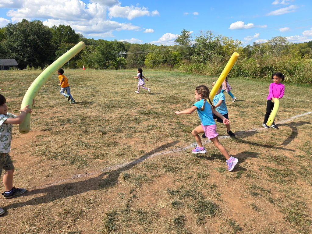 Children run across a grassy field holding foam pool noodles, using them as playful taggers during a group activity.