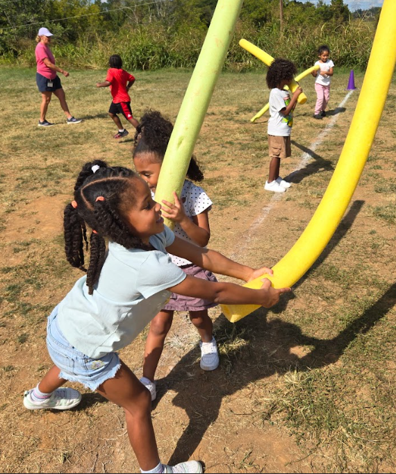 Children run across a grassy field holding foam pool noodles, using them as playful taggers during a group activity.